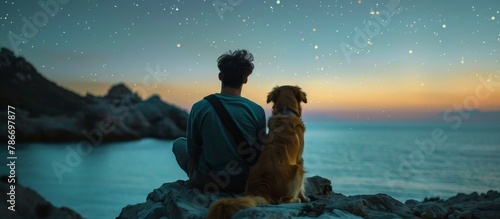 Young man with golden retriever admiring starry night sky on sea coast, sitting together, rear view
