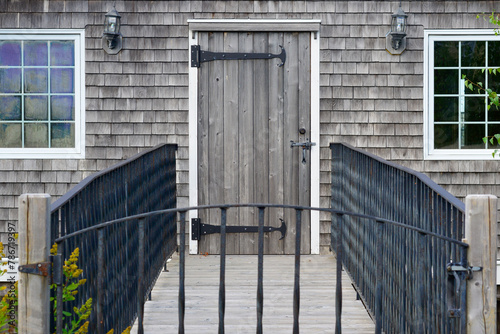 The entrance to a grey weathered worn boathouse with a solid wood door, black metal hinges, and white trim. There are two glass windows and a bridge leads to the door with a black handrail and gate.