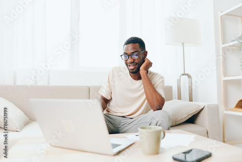 Happy African American man sitting on a modern sofa in a cozy living room, typing on his laptop He is a smiling freelancer working from home, enjoying the comfort and convenience of cyberspace