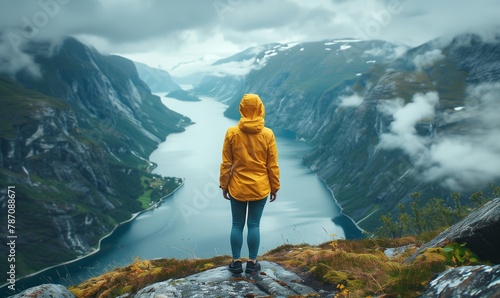 Portrait of a woman in a yellow raincoat standing from her back on a troll's tongue high in the mountains of Norway against the background of fjords