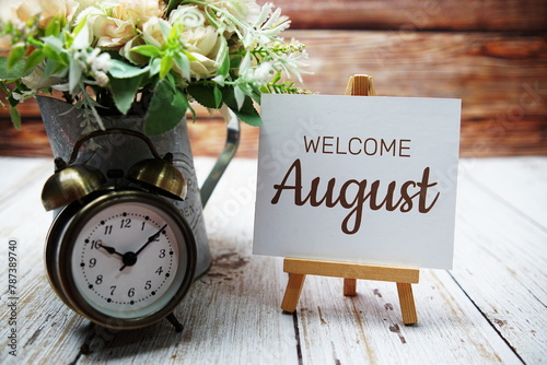 Welcome August text message written on paper card with wooden easel and alarm clock with flower in metal vase decoration