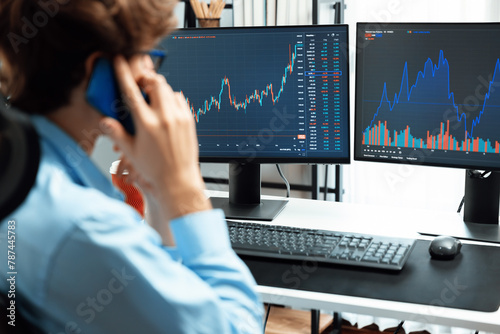 Young investor stock trader calling on smartphone to broker, discussing with dynamic financial exchange investment on two laptop screens in current market graph in real time at modern office. Gusher.