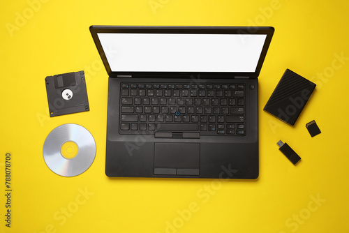 Evolution of storage media. Laptop with CD disk, sd card, floppy disk, USB flash drive and external hard drive on yellow background. Flat lay