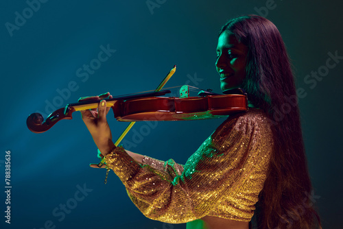 Cheerful young Indian woman, violinist in festive attire performing in neon light against gradient background. Concept of art, music, hobby, classical music and modern lifestyle. Ad
