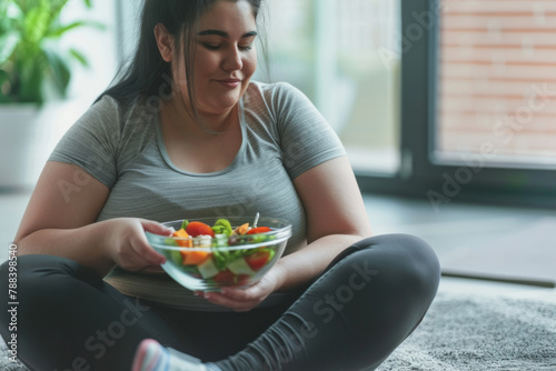 A plump Arab woman in athletic clothes savors a vegetable salad from a glass bowl while seated on the floor, committed to following a slimming and exercise program.
