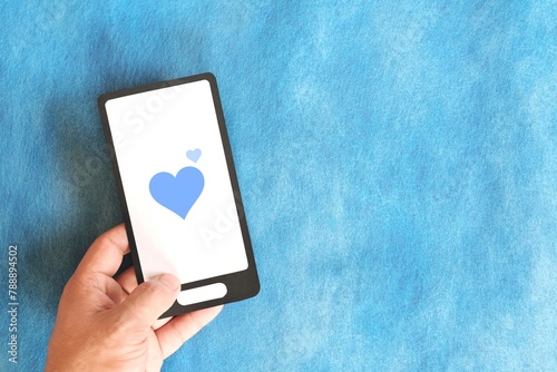 Human hand holding mobile phone with blue heart icon. Kindness, love and positivity in social media internet concept.