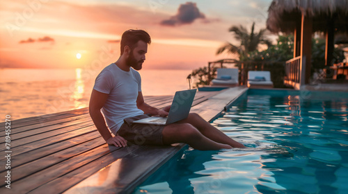 Young man sitting poolside by the swimming pool working on laptop notebook computer during sunset. Tropical island summer vacation or holiday remote online work on the internet, freelancer