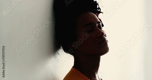 One pensive young black woman with eyes closed leans on wall with thoughtful contemplative expression. 20s Brazilian person of African descent in quiet solitude