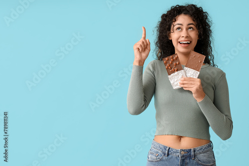 Beautiful young African-American woman with sweet chocolate bars pointing at something on blue background