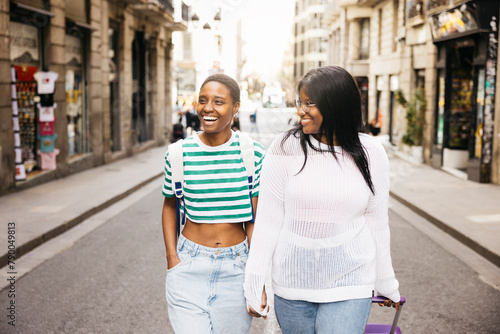 A young female couple is exploring a new city during a short weekend trip, walking together while pulling a suitcase along a city street.