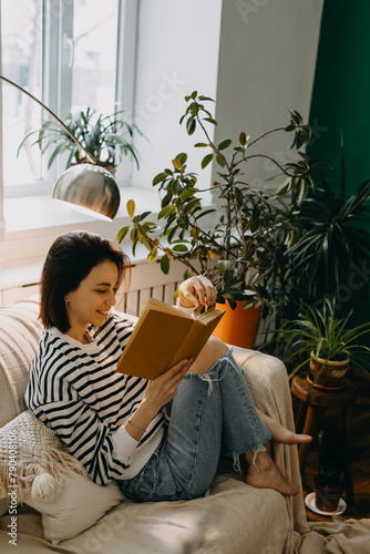 Young happy woman reading a book on a couch, in soft day light by a window, surrounded by green houseplants.