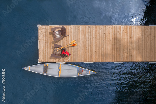 Aerial view two Adirondack chairs on a wooden dock by a serene Ontario lake on a sunny summer morning. A yellow canoe is tied nearby, with visible life jackets and oars.