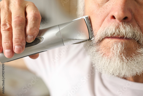 Man trimming beard with electric trimmer indoors, closeup