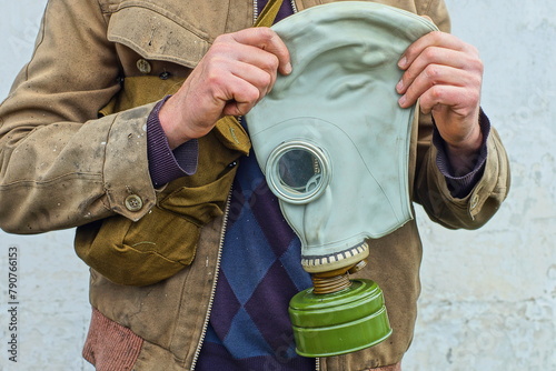 one military gray rubber protective gas mask with a green iron filter in the hands of a man dressed in a dirty yellow jacket near a white wall during the day on the street