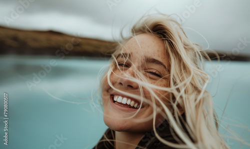 Close up photo of a blonde woman smiling and looking at the sky, taking a selfie at the Icelandic blue lagoon with her white hair blowing in the wind