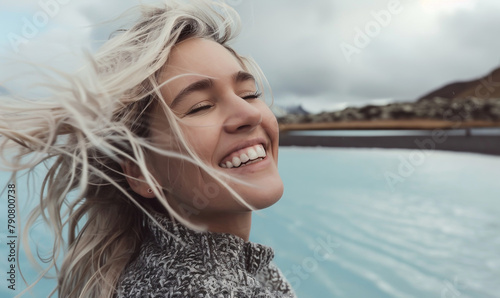 Close up photo of a blonde woman smiling and looking at the sky, taking a selfie at the Icelandic blue lagoon with her white hair blowing in the wind