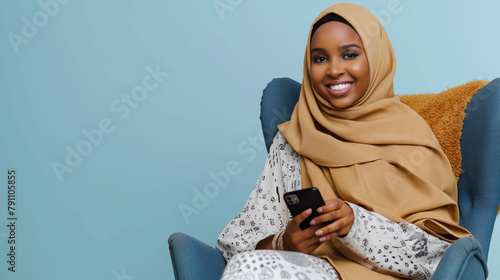 Happy African Muslim woman in abaya hijab using mobile phone while sitting on the armchair and looking at the camera on isolated blue background with space for copy