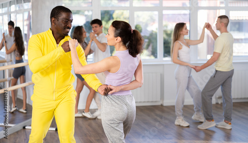 Positive smiling young african american man and female partner dancing cha-cha-cha with group of guys and girls in modern dance class