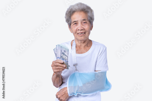 A sen woman with broken arm and wearing a sling with a smiling face and holding dollars for insurance and paid care isolated on a white background. Old elderly female medical care. Health care concept