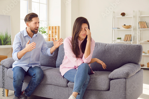 Angry husband shouting in quarrel on his unhappy upset wife sitting on sofa at home. Young couple quarreling and arguing in living room. Marriage problems, relationship and divorce concept.