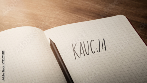 A handwritten inscription "Kaucja" on a grille of an open notebook on a wooden countertop, next to a black pencil, lighting of light. (selective focus), translation: deposit