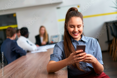 Smiling asian businesswoman in smart casual wear uses smartphone, team works at wooden table in modern office. Woman checks app, coworkers discuss project in blurred background, communication.