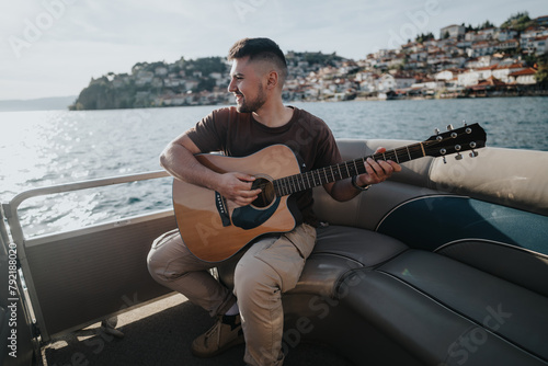 Casual guitarist smiles as he enjoys music on a sunny day with the ocean and cityscape behind him.