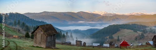 View of mountain village surrounded by peaks of Carpatian mountains with wooden cabins and faint trail of an automobile tires on dews grass. Mist gently blankets mountain valley village in early hours