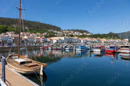 A serene marina of Muros in Spain with various boats docked in calm waters, with quaint hillside houses and clear blue sky in the background