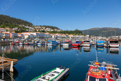 A serene marina of Muros in Spain with various boats docked in calm waters, with quaint hillside houses and clear blue sky in the background