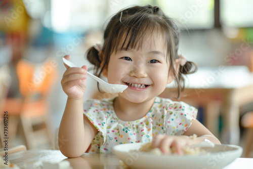 a happy young Asian girl eating at a table in a children's restaurant, holding a spoon and plate, with long hair bangs, focusing on her face, in natural light