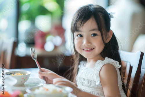 a happy young Asian girl eating at a table in a children's restaurant, holding a spoon and plate, with long hair bangs, focusing on her face, in natural light