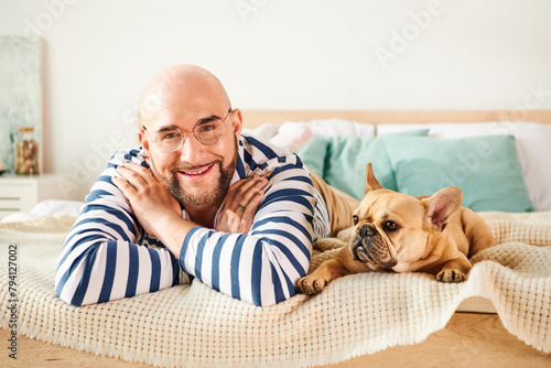 Handsome man with glasses relaxing on bed next to his loyal French Bulldog.