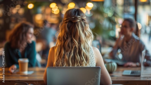 A young woman types away on a laptop back to the camera while a group of friends sit together at a nearby table sharing stories . .