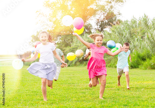 Portrait of cheerful preteen boys and girls with colorful toy balloons in hands running on green lawn in city park on sunny summer day