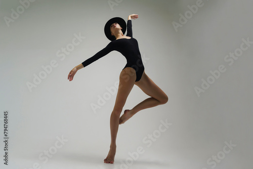 young ballerina in a black bodysuit shows ballet steps in motion standing on one leg and spreading her hands in a hat