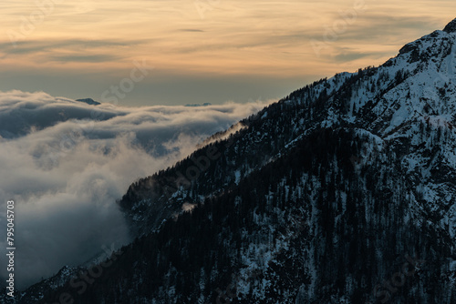 visuale dettagliata e da lontano di un'immensa quantità di nuvole basse che avanzano tra le valli in mezzo alle montagne innevate, in inverno, al tramonto, con un cielo colorato di arancione