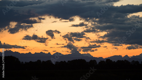 visuale in primo piano di un cielo nuvoloso durante un tramonto color arancione e delle sagome scure delle montagne distanti viste da lontano 