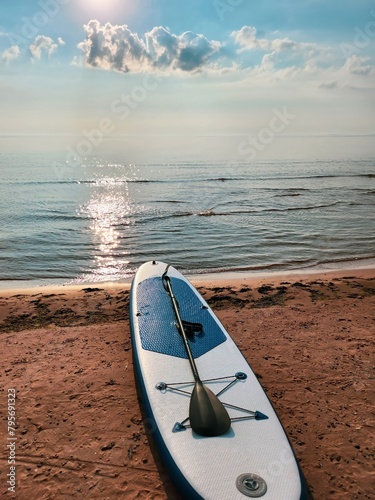 Blue and white paddleboard on sandy beach by calm lake under cloudy sky with sunbeams in morning. Vertical orientation with copy space, ideal for water sports or summer blog.