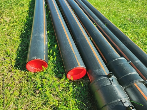An assortment of black plastic pipes of varying diameters arranged on lush green grass, creating an industrial scene against a vivid green backdrop, harmonizing utility with nature
