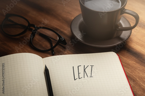  A handwritten inscription "Leki" on a grille of an open notebook on a wooden countertop, next to a black pencil, a cup with coffee and glasses, a flash of light. (selective focus)