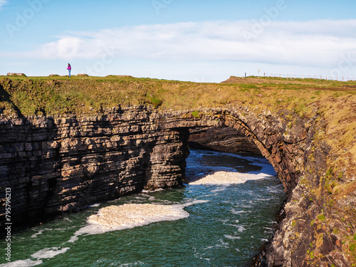 Tourist enjoy stunning Bridges of Ross in county Clare, Ireland. Rock formation arched over ocean creating natural bridge. Popular tourist landmark and point of interest. Warm sunny day.