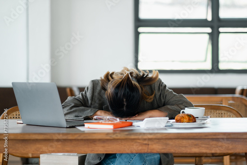 Overworked individual takes a quick nap at their desk, surrounded by a laptop, papers, and a half-eaten breakfast, depicting workplace fatigue.