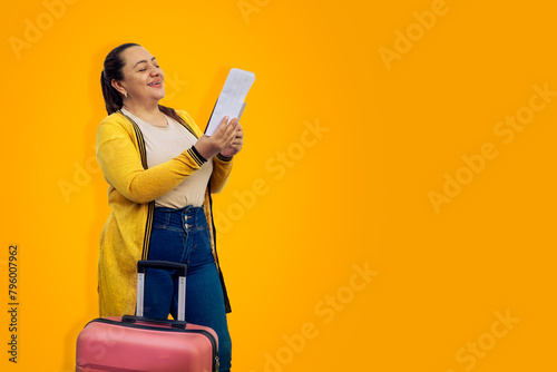 Beautiful woman in summer clothes with pink suitcase, happy with her hand up, celebrating her cheap ticket on yellow background. Tourist very happy to travel abroad with a bargain ticket. Copy space.