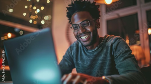 Joyful African-American man wearing glasses working on his laptop in a warmly lit room.