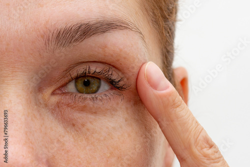 Close up cropped eye of middle aged caucasian woman looking at camera holding finger on eyelid