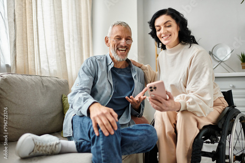 A man sits next to his wife on a wheelchair with smartphone in a cozy living room.