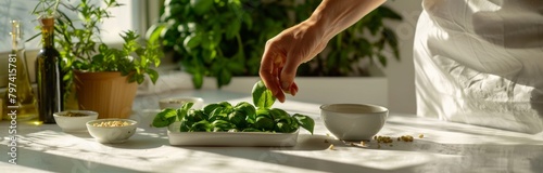 a person preparing basil on a white table, with ingredients like pine nuts and olive oil in small bowls beside them
