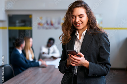 Businesswoman in smart casual attire smiles at smartphone in office setting, team discussion blurred in background. Pro multitasking, connects online, stays informed with tech corporate environment.