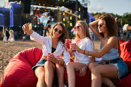 Group of women enjoy music fest on beach, lounging on bean bags. Casual attire, sunglasses, sipping drinks, one takes selfie. Warm daylight highlights relaxed, fun atmosphere of outdoor summer event.